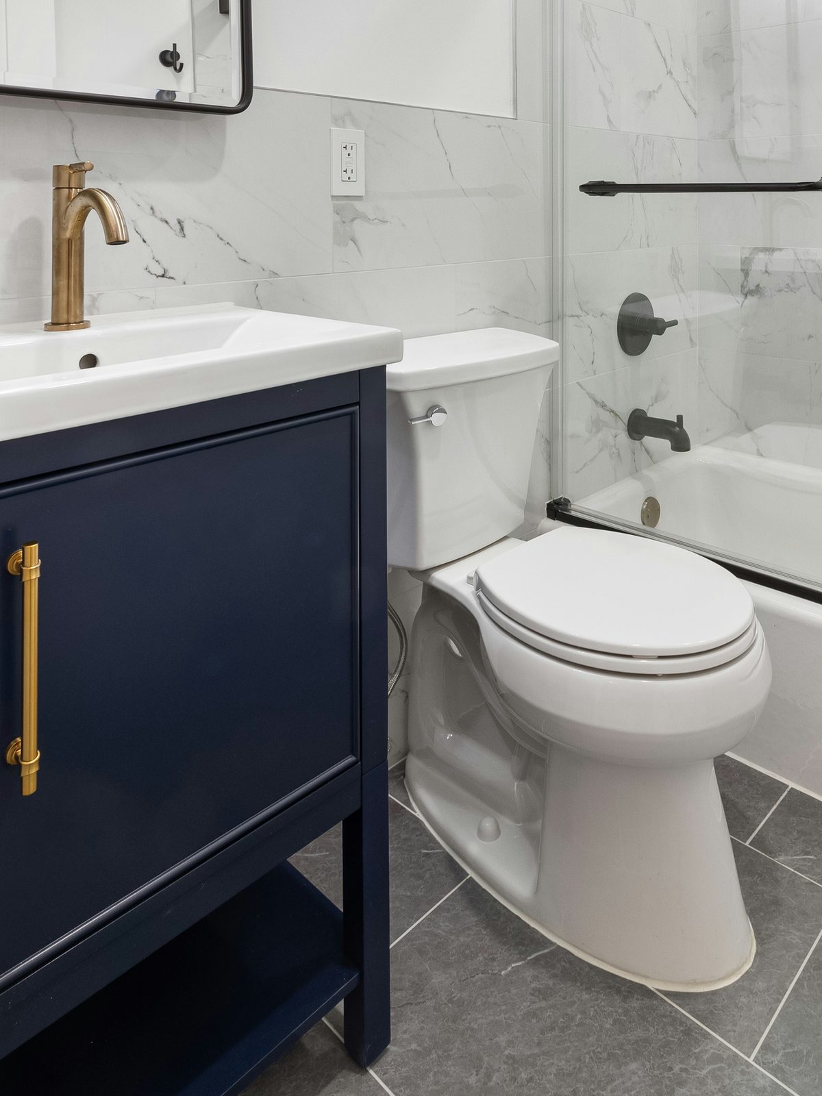Bathroom with a navy vanity, brass faucet, white toilet, marble-look wall tile, and a tub with a sliding glass door