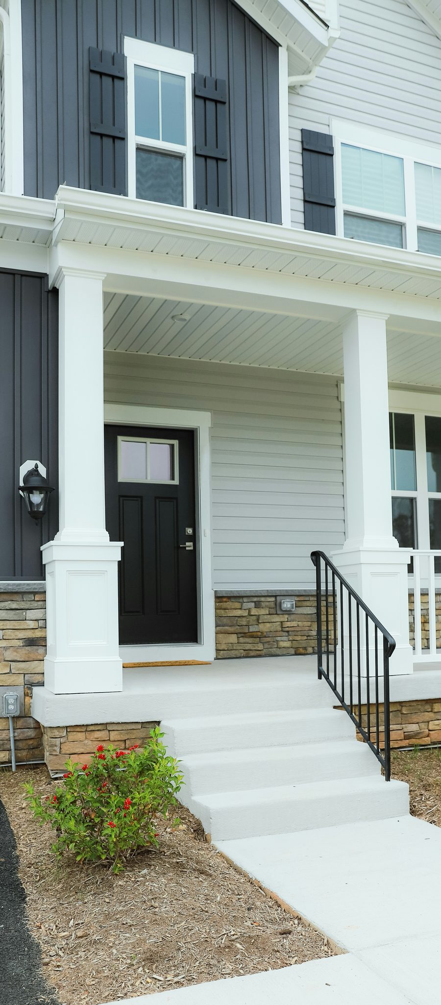 Covered front porch with white columns, a black front door, stone veneer, and concrete steps with a black railing